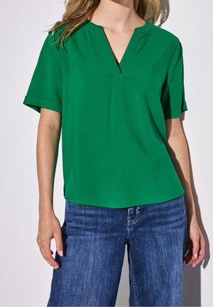 Vihreä V-kaula-aukkoinen paita lyhyillä käännetyillä hihoilla, sileä kangas, rento istuvuus ja kaareva helma. Yhdistetty sinisiin denimlaatuisiin leveälahkeisiin farkkuihin.