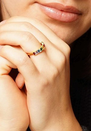 Goldring mit farbigen Edelsteinen in einem geschwungenen Design. Glatte Metalloberfläche mit rechteckigen und quadratischen, rot, grün und blau geschliffenen Steinen.