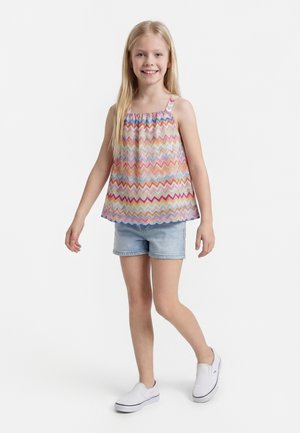 Χαμογελαστή κοπέλα με μακριά ξανθά μαλλιά, ντυμένη με πολύχρωμο αμάνικο τοπ με ζιγκ-ζαγκ μοτίβο, τζιν σορτς και λευκά παπούτσια χωρίς κορδόνια, στέκεται σε λευκό φόντο.