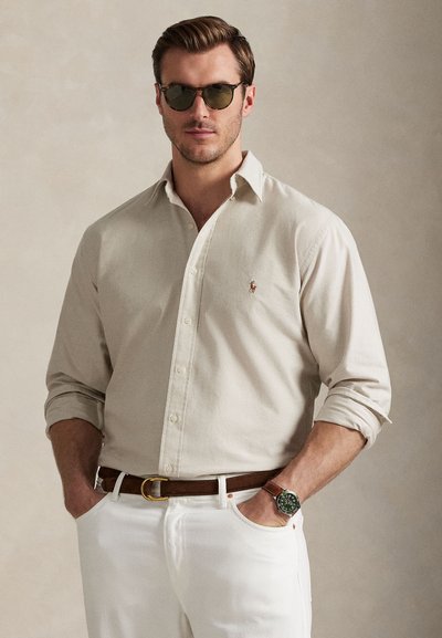 Camicia beige con bottoni e maniche arrotolate, pantaloni bianchi, cintura marrone e orologio verde. Il modello indossa occhiali da sole scuri e posa davanti a uno sfondo neutro.