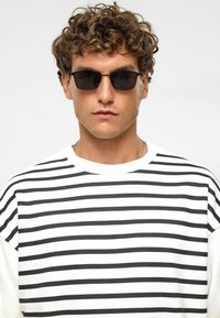 Zwart-wit gestreept t-shirt, ontspannen pasvorm, gedragen met rechthoekige donkere zonnebril. Gemêleerde stof zichtbaar in close-up.