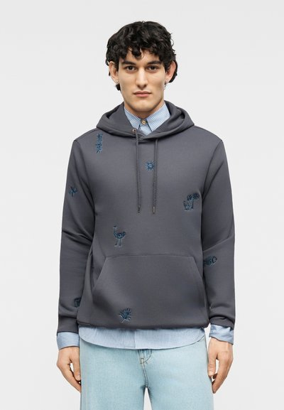 Jeune homme portant un sweat à capuche gris avec des motifs brodés d'animaux et de plantes bleus, une chemise à rayures bleu clair et un jean délavé clair.