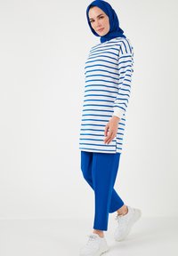 Túnica de rayas azules y blancas con un corte largo y aberturas laterales, combinada con pantalones tapered de color azul sólido y zapatillas deportivas blancas.