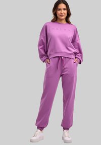 Lawendowy dwuczęściowy zestaw do noszenia w domu, składający się z luźnej bluzy z okrągłym dekoltem i haftowanym logo oraz spodni z elastycznymi mankietami.