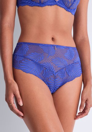 Femme portant une culotte taille haute en dentelle bleue et un soutien-gorge assorti, debout, les mains relâchées le long du corps sur fond uni.