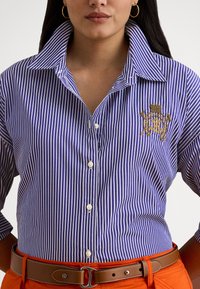 Žena oblečená v modro-bielej zvislo pruhovanej košeli na gombíky s vyšívaným erbom a hnedým opaskom cez oranžové nohavice.