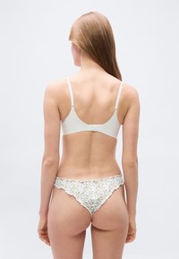 Femme aux longs cheveux portant un soutien-gorge blanc et une culotte bikini à motif floral, vue de dos sur un fond uni.
