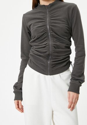 Femme portant une veste zippée gris foncé froncée avec des poignets côtelés et un pantalon blanc ample sur un fond uni.