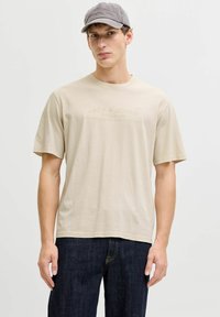 Beiges Baumwoll-T-Shirt mit kurzen Ärmeln und einem dezenten Logodruck. Das Modell kombiniert es mit dunklen Denim-Jeans und einer grauen Mütze.