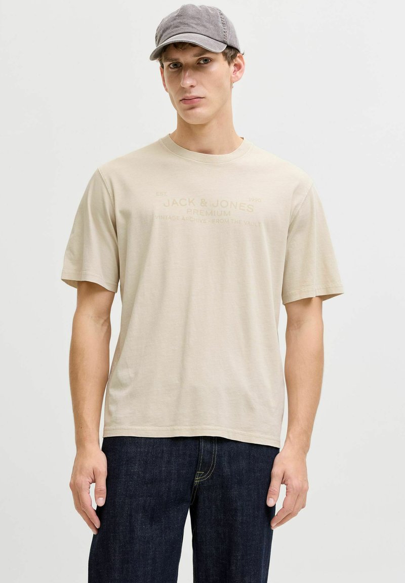 Beiges Baumwoll-T-Shirt mit kurzen Ärmeln und einem dezenten Logodruck. Das Modell kombiniert es mit dunklen Denim-Jeans und einer grauen Mütze.