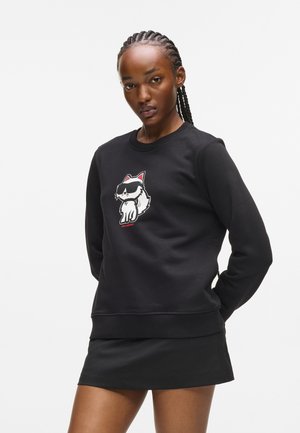 Zwarte sweatshirts met een cartoonkat in een zonnebril, ronde hals, ribgebreide manchetten en tailleband. Gecombineerd met een zwarte minirok.