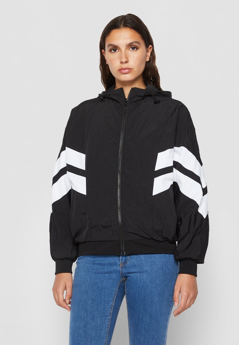 Naine kannab musta tõmblukuga kapuutsiga jopet, mille varrukatel on valged nurgelised triibud, ja siniseid teksapükse, seisab tavalise heleda tausta ees.