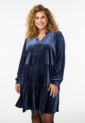 Mujer sonriente con cabello rizado, vistiendo un vestido de terciopelo azul marino de mangas largas y con volantes, contra un fondo blanco liso.