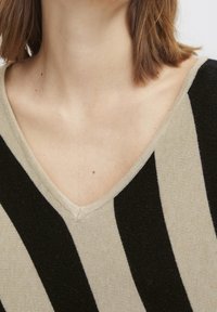 Beżowy i czarny sweter w paski z dekoltem w kształcie V. Tkanina wydaje się miękka o gładkiej fakturze i kontrastujących, intensywnych paskach.