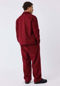 Completo giacca e pantaloni rossi dalla vestibilità rilassata, caratterizzato da una texture liscia, ricamo sottile sulla schiena e abbinato a calzature nere.