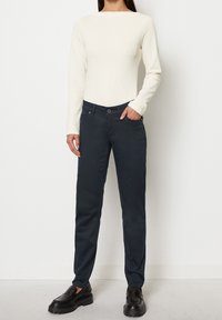Maglietta bianca a maniche lunghe e pantaloni blu navy slim-fit con un design a cinque tasche. Scarpe nere slip-on con suola spessa completano il look.