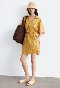Femme portant une robe jaune à fleurs à manches courtes, un chapeau bob beige, des sandales blanches à enfiler et portant un grand sac fourre-tout tressé marron foncé.