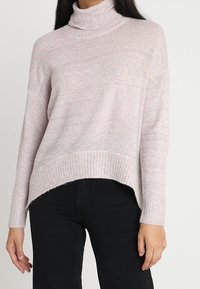 Hellrosa Rollkragenpullover aus weichem Material, mit geripptem Saum und langen Ärmeln, leicht übergroß geschnitten und mit einem High-Low-Design.
