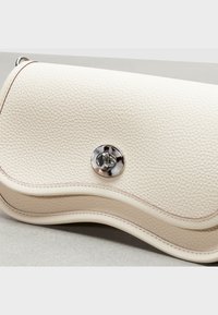 Kremowa skórzana torebka o fakturowanej powierzchni, zaokrąglonym kształcie, kontrastującym przeszyciu oraz srebrnym zapięciu z przodu.