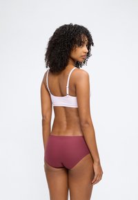 Γυναίκα με μπούκλες, που φοράει ανοιχτό ροζ σουτιέν και μπεζ εσώρουχα χωρίς ραφές σε μπορντό χρώμα, στέκεται με την πλάτη προς την κάμερα σε λευκό φόντο.