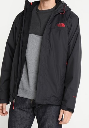 Osoba nosząca czarną kurtkę North Face z czerwonym logo, szarą bluzę i ciemne dżinsy, stojąca na jednolitym tle.
