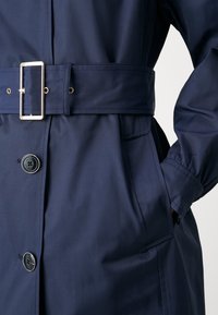 Granatowy trencz z paskiem w talii, dużymi guzikami, bocznymi kieszeniami i elastycznymi mankietami, wykonany z gładkiej, lekkiej tkaniny.