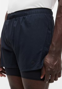 Μπλε ναυτικό αθλητικό σορτς με ελαστική μέση, εσωτερική μαύρη φόδρα και διακριτική λεπτομέρεια λογοτύπου στο τελείωμα, κατασκευασμένο από ελαφρύ ύφασμα.