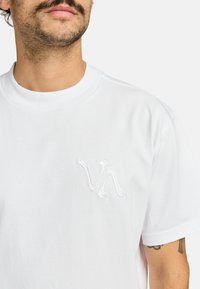 Weißes Baumwoll-T-Shirt mit einem kleinen gestickten Logo auf der linken Seite. Das Design umfasst einen Rundhalsauschnitt und kurze Ärmel.