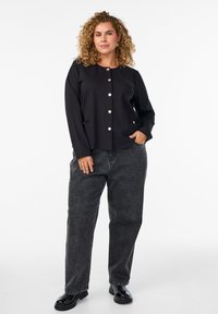 Svart långärmad jacka med rund halsringning och stort knäppning, kombinerad med grå vida jeans och svarta blanka skor.