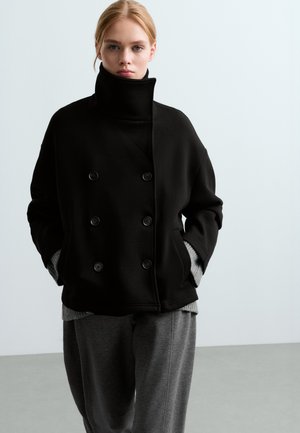 Une femme porte un manteau noir croisé à col montant et un pantalon gris devant un fond de studio uni.