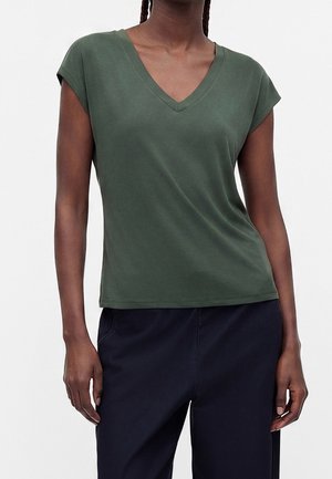 Vrouw draagt een donkergroene V-hals top met korte mouwen en een zwarte broek, staand tegen een effen lichte achtergrond.