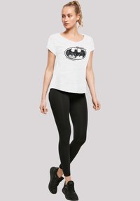Valkoinen lyhythihainen t-paita, jossa on musta Batman-logo, yhdistettynä mustiin legginsseihin ja mustiin urheilukenkään. Sujuva kangas, rento malli.