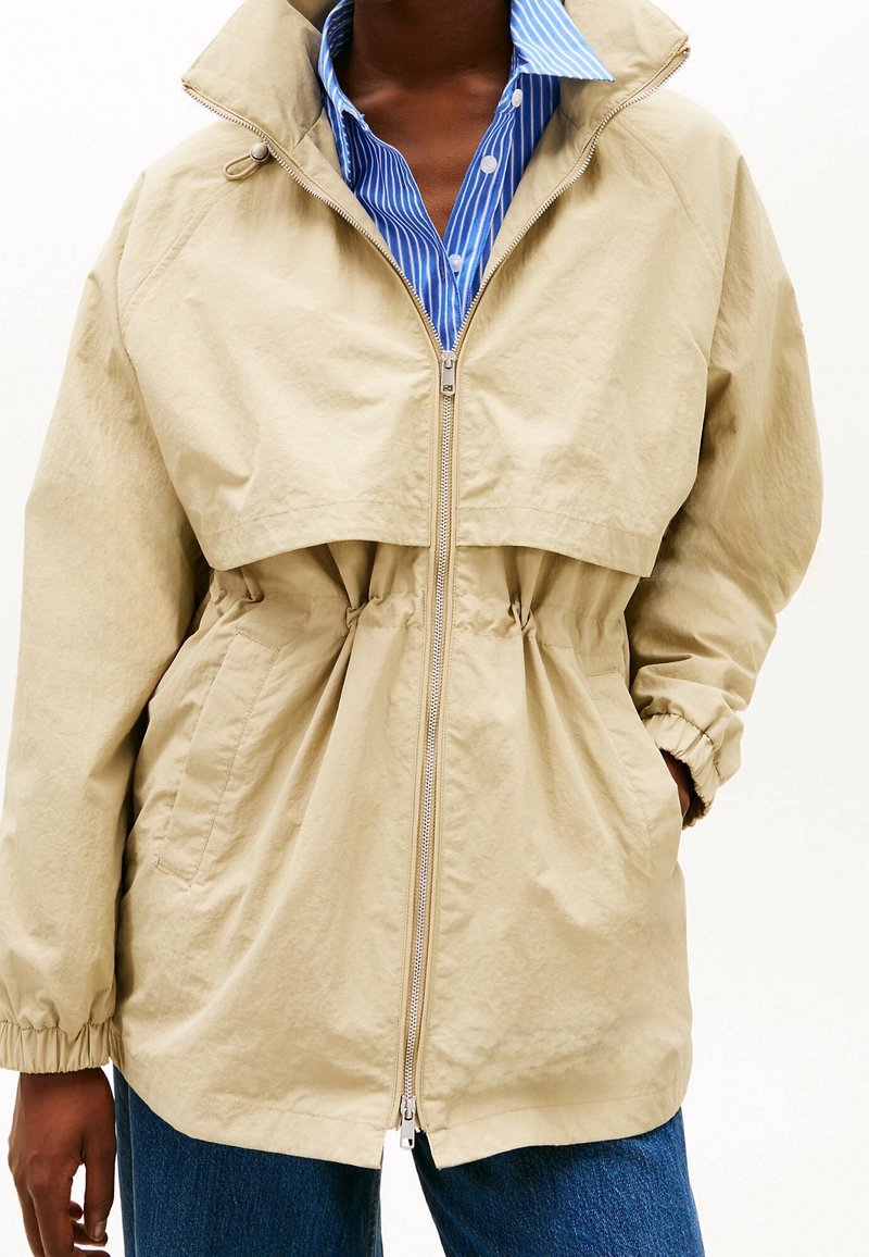 Personne portant une veste beige zippée sur une chemise à rayures bleues et blanches, les mains dans les poches de la veste et un jean bleu visible.