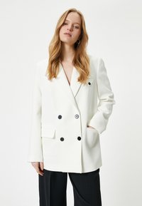 Hvid dobbeltradet blazer med klapkrave og sorte knapper. Har en lomme på venstre bryst og en sidelomme. Glat, struktureret stof.