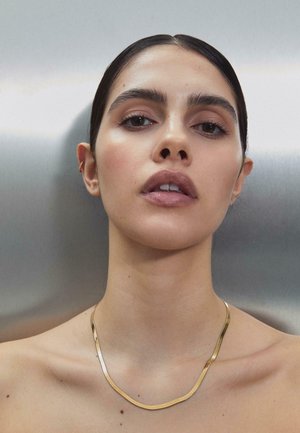 Nainen, jolla on sileä, tumma hius kammattuna taakse, pukeutunut ohueen kultakaulakoruun ja pieneen korvan koruun, yksinkertaista harmaata taustaa vasten.