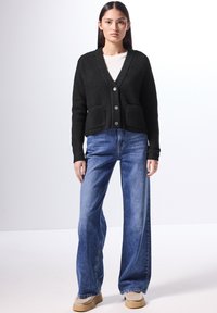 Svart stickad cardigan med V-ringning, tre knappar och två fickor fram, matchad med vida ljusblå jeans och beigefärgade skor.