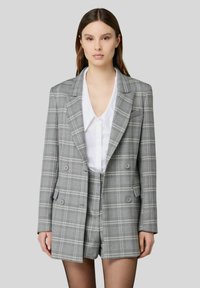 Blazer grigio a quadri con bottoni doppi, abbinato a pantaloni corti coordinati. Presenta un colletto a punte e una camicia bianca sotto.