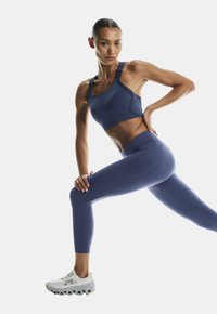 Marineblaue sportliche Sport-BH mit verstellbaren Trägern, kombiniert mit hochtaillierten marineblauen Leggings. Weiße Laufschuhe mit strukturierten Sohlen.