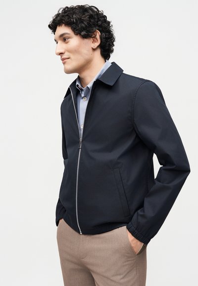 Άνδρας με σγουρά μαλλιά, φορώντας μπλε ζακέτα με φερμουάρ πάνω από ανοιχτό μπλε πουκάμισο και μπεζ παντελόνι, με τα χέρια στις τσέπες, στραμμένος αριστερά σε απλό φόντο.