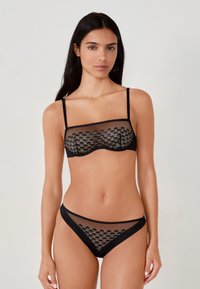 Conjunto de lencería negra que incluye tela de malla transparente, sujetador estructurado con detalles estampados y braguitas de tiro bajo a juego con acentos similares.