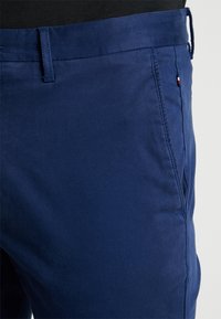 Närbild av marinblå byxor med bältesöglor, sidoficka och en liten tagg i rött, vitt och svart på fickans söm.