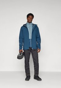 Sinine veekindel kapuutsiga jakk, kandes selle all helesinist sisejakki, mustad püksid ja mustad kingad. Käes kaitsmet kiiver.