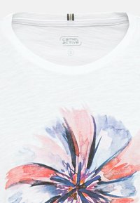 T-shirt blanc avec un motif floral dans des tons de rose, violet et bleu. Encolure ronde avec une texture subtile, étiquette visible à l'intérieur du col.
