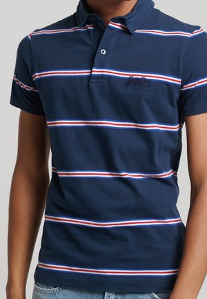Person iført en mørkeblå polo med hvide og røde vandrette striber og lyseblå jeans, vist fra nakken til hofterne.