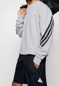 Man med grå sweatshirt med tre svarta ränder på ärmarna och svarta Adidas-shorts, som håller en svart väsksnodd över axeln.