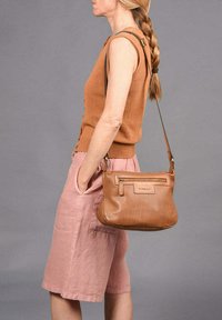 Tan geperforeerde leren handtas met een ritszak, een ronde vorm en een verstelbare schouderband, gedragen als crossbody over lichtroze shorts.