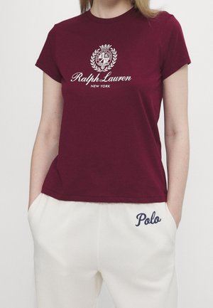Kobieta w bordowej koszulce Ralph Lauren z logo i białych spodniach z napisem "Polo" na kieszeni.