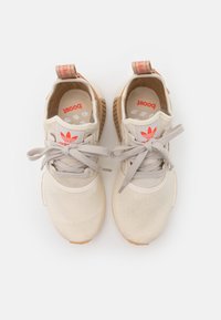 Beiga sportskor med en texturerad tygöverdel, ljusgrå snören och en sula av gummirubber. Har röda detaljer på hälen och tungan.