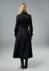 Czarna satynowa bluzka z długimi rękawami i wysokim kołnierzem, zestawiona z ciemną spódnicą z plisami, złotymi guzikami i zapięciem na zamek z tyłu.