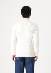 Witte kabelbrei sweater met hoge kraag, geribbelde manchetten en zoom; gecombineerd met blauwe denim jeans, die een klassieke textuurontwerp laten zien.